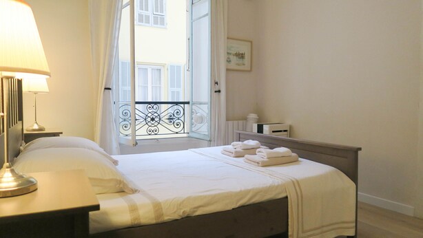 A neatly made bed with white linens and towels, a bedside lamp, a small bedside table, and a view of a balcony with a railing. - Family-friendly apartment in an authentic neighborhood and its open market (Nice)