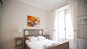 A bedroom with a wooden bed, white bedding, two bedside tables with lamps, a painting on the wall, and a window with curtains. - Family-friendly apartment in an authentic neighborhood and its open market (Nice)