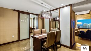 A modern hotel room with a dining area featuring a wooden table and leather chairs, a city view, and a separate seating area with a sofa and armchairs.