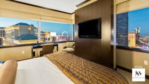 A hotel room with a large bed, a TV mounted on the wall, and a view of the cityscape through large windows.