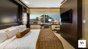 A hotel room with a large bed, a desk, a chair, a TV, and a view of a cityscape.
