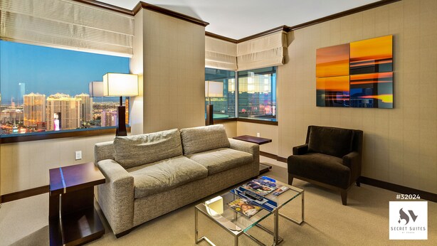 A modern hotel room with a city view, a sofa, a chair, a coffee table, and a painting on the wall.