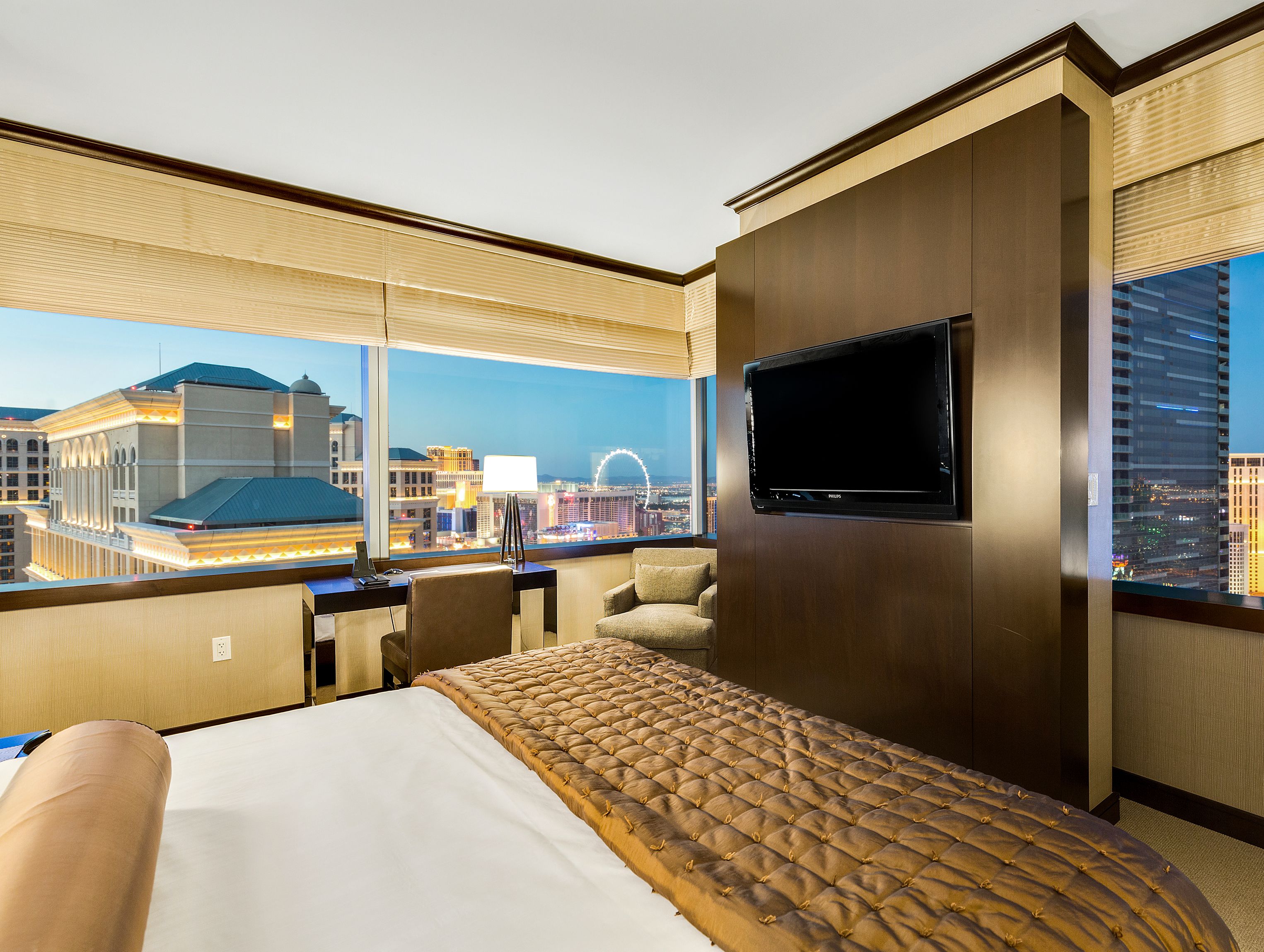 Un cuarto de hotel con una cama grande, un televisor montado en la pared y vistas de la ciudad a través de amplios ventanales.