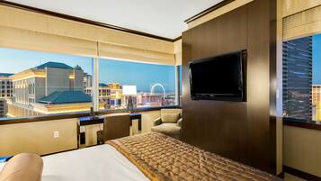 Un cuarto de hotel con una cama grande, un televisor montado en la pared y vistas de la ciudad a través de amplios ventanales.