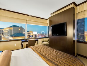 Ein Hotelzimmer mit einem großen Bett, einem an der Wand montierten Fernseher und Blick auf die Stadt durch große Fenster.