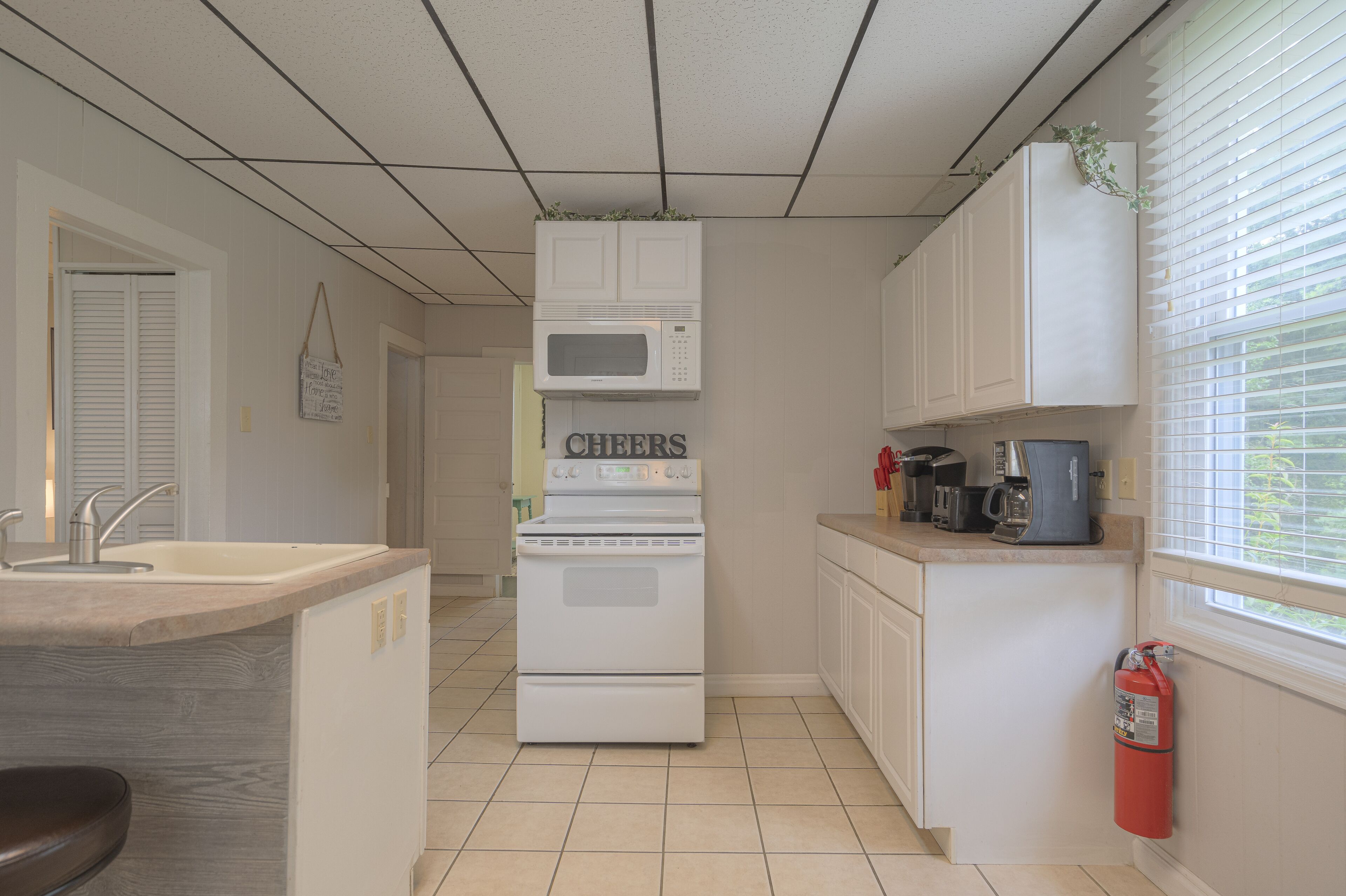Een compacte keuken met een ingebouwde magnetron, fornuis en spoelbak. Een brandblusser is aan de muur, bij het raam, bevestigd.