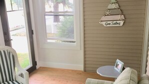 A room with a green and white striped sofa, a wicker chair, a small round table with a picture frame, a window with a view of greenery, and a sign on the wall that says "Gone Sailing". - 
VENETIAN'27SEND INQUIRY! Walk to Everything/Fenced Yard/Enclosed Front Porch/AC (Charlevoix)