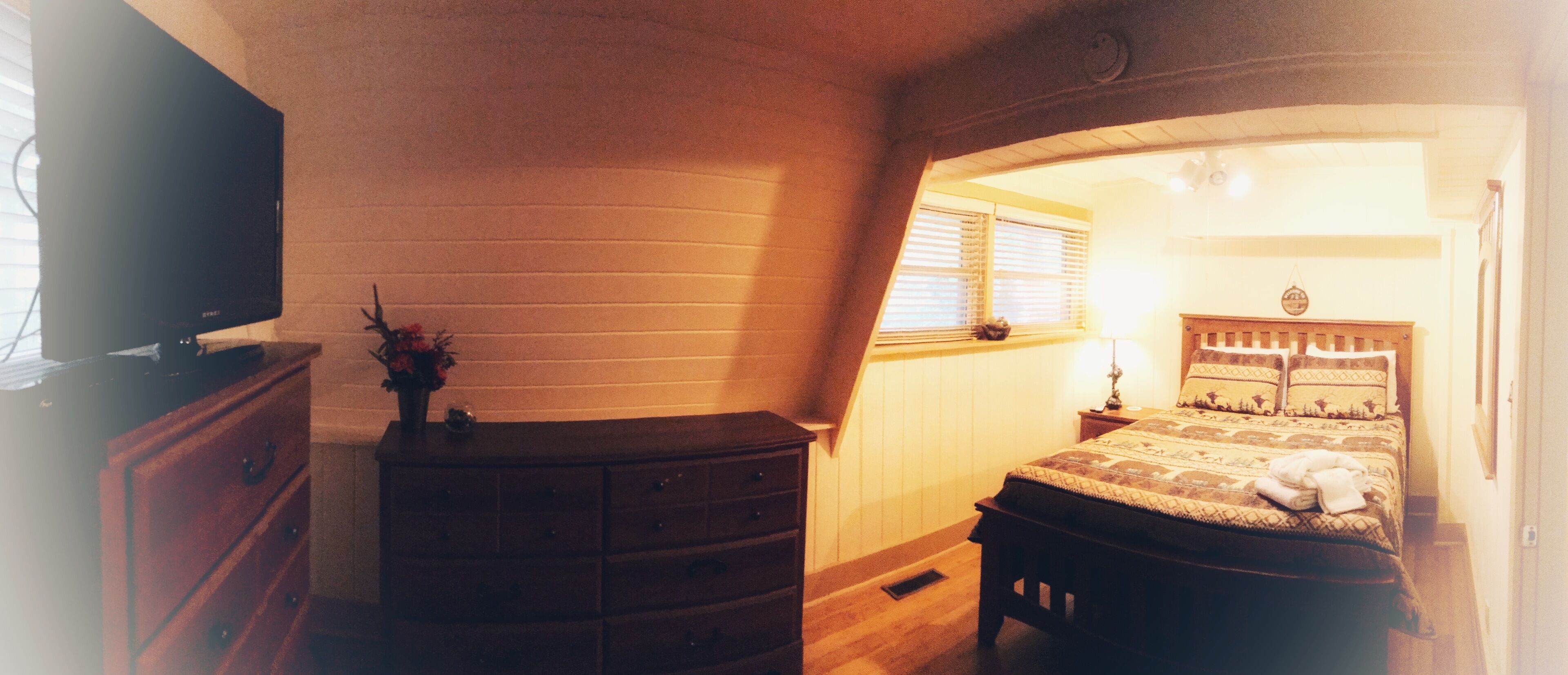 Quarto com cômoda de madeira, cama com roupa de cama estampada, janela com persianas e mesa de cabeceira com abajur.