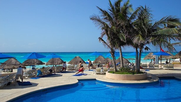 Zona de piscina con sillas de descanso y sombrillas, una palmera y vistas al océano.