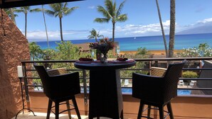 A balcony with a table and chairs, overlooking a beach with palm trees and the ocean. - Amazing panoramic ocean view!   Perfect for couples. Just steps to the ocean.🏝 (Lahaina)