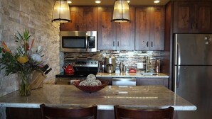 A kitchen with wooden cabinets, a stainless steel refrigerator, a microwave, and a large island with chairs. - Amazing panoramic ocean view!   Perfect for couples. Just steps to the ocean.🏝 (Lahaina)