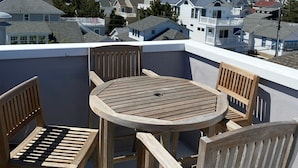 Une terrasse en bois avec une table ronde et quatre chaises, donnant sur un quartier résidentiel avec des maisons et un ciel dégagé.