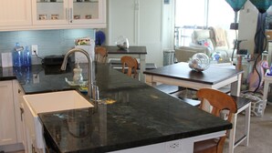 Una cocina moderna con armarios blancos, encimera de granito negro y una isla integrada con fregadero. Se observan sillas de madera y una mesa de comedor al fondo.