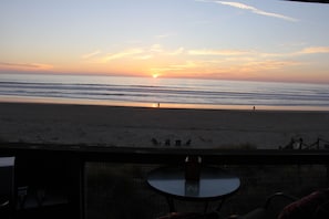 Une vue sur la plage depuis un balcon, avec une table et des chaises, donnant sur l’océan et un coucher de soleil.