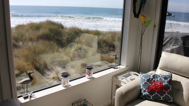 Una habitación con un amplio ventanal con vistas al océano, vista a la playa, una pequeña mesa con una lámpara y un sillón con un cojín estampado.