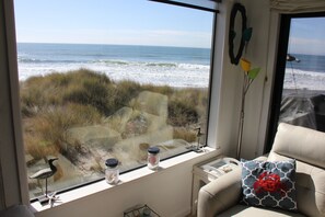Une pièce dotée d’une grande fenêtre donnant sur l’océan, avec vue sur la plage, une petite table avec une lampe et un fauteuil confortable agrémenté d’un coussin à motifs.