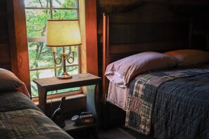 A bedroom with a wooden headboard, a bedside table with a lamp, and a window with a view of greenery.
