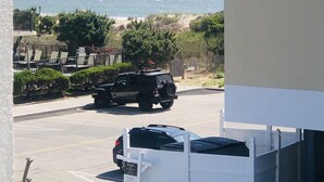 Un aparcamiento con una camioneta SUV negra y un coche blanco, un edificio blanco con balcones, y vistas a la playa y al océano.