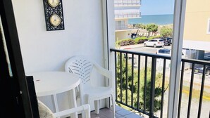 Un balcón con una silla blanca y una mesita, un tapete azul, y vistas al mar y edificios.