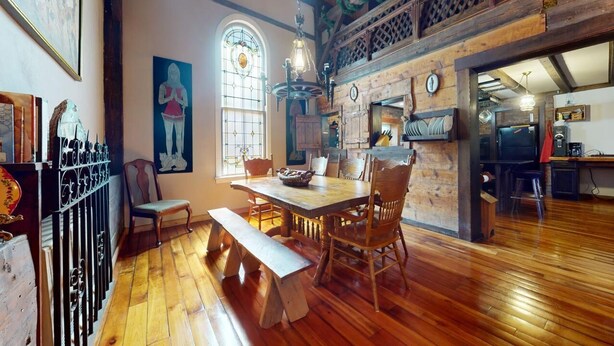 Un coin repas rustique avec un plancher en bois, une grande table et des chaises. On y trouve également un banc, un vitrail et un tableau accroché au mur.