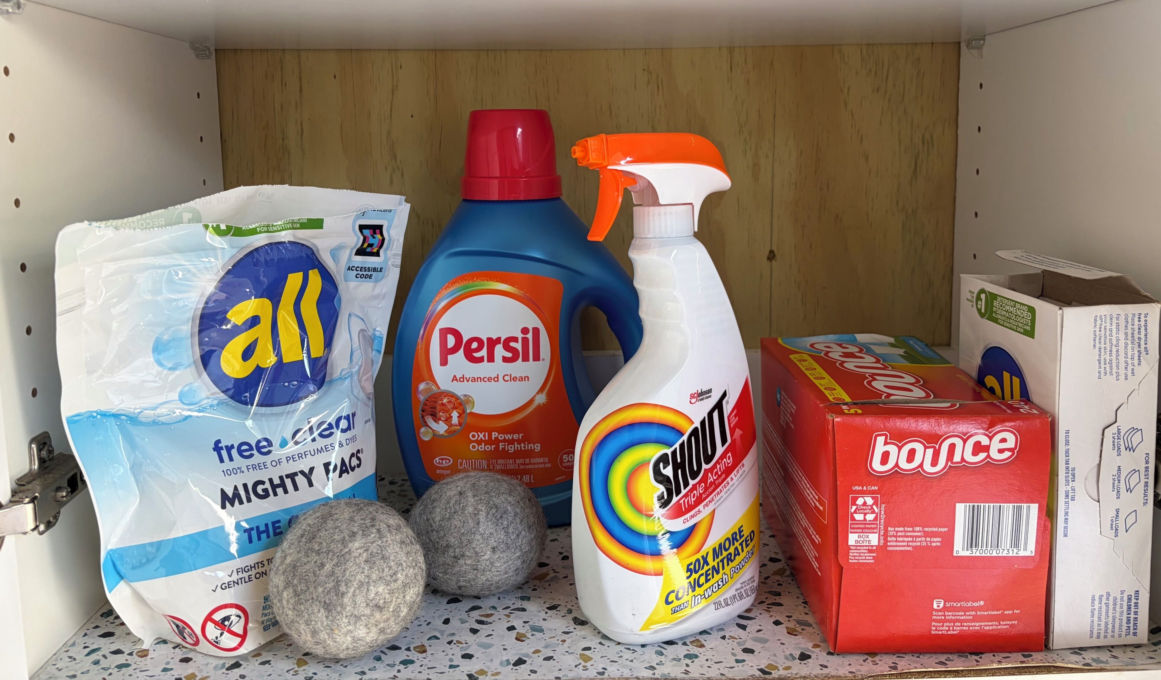 Een plank met schoonmaakmiddelen, waaronder een zak 'all' schoonmaakproducten, een fles 'Persil', een 'Swim' wasmiddel en een doos 'Bounce'.