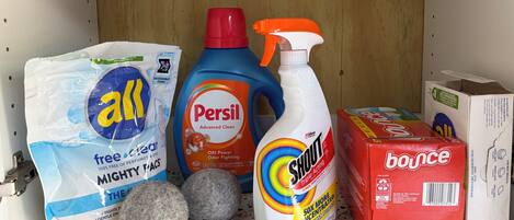 Estante con productos de limpieza, incluyendo un paquete de productos de limpieza varios, una botella de 'Persil', detergente 'Swim' y una caja de 'Bounce'.