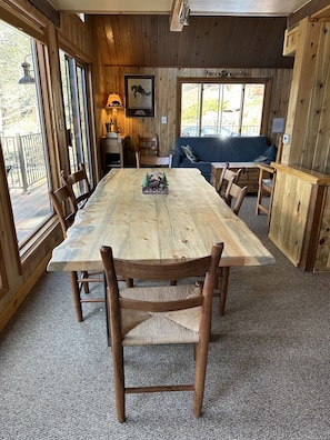 Una mesa de comedor de madera con sillas, un adorno pequeño y un sofá azul en una habitación de estilo cabaña con ventanas grandes y techo de madera.