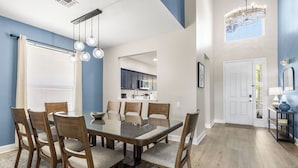 A dining area with a glass-top table and wooden chairs, a kitchen in the background, and a door leading to another room. - Welcome to The Windsor Magic with Professional Arcade Room! Brand New 2024! (Kissimmee)