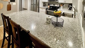 Une cuisine dotée de plans de travail en granit, d’un îlot central avec des tabourets de bar, d’appareils électroménagers en acier inoxydable et de luminaires suspendus.