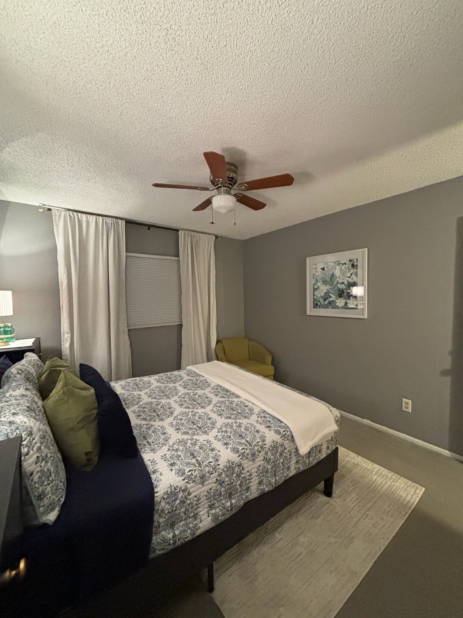 Une chambre à coucher avec un lit, un ventilateur de plafond, une chaise, une lampe et un tableau au mur.