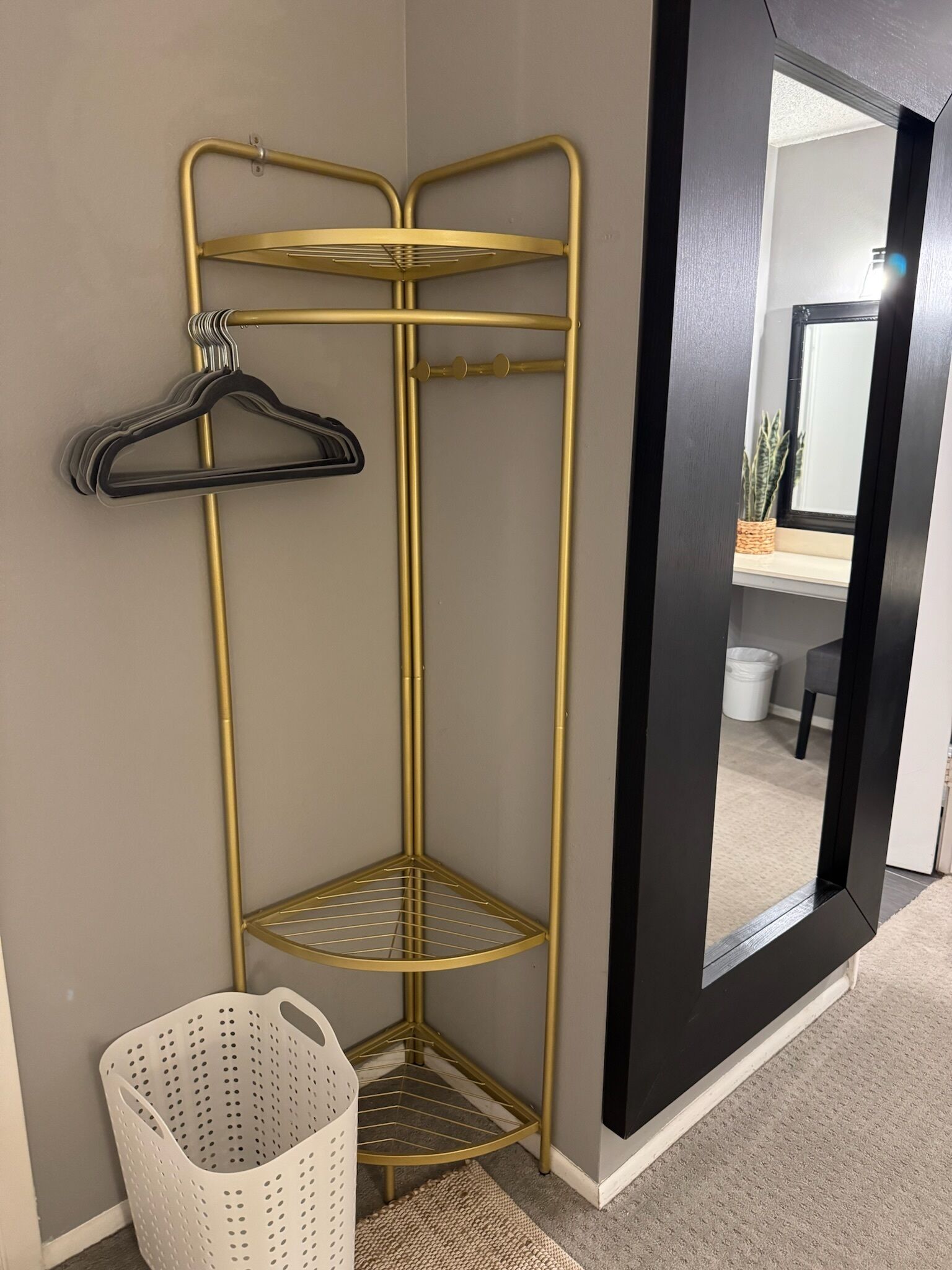 Un meuble d’angle pour le rangement, doté de plusieurs étagères et d’une tringle pour suspendre des vêtements, un panier à linge blanc et un miroir qui reflète une pièce avec une plante et une chaise.