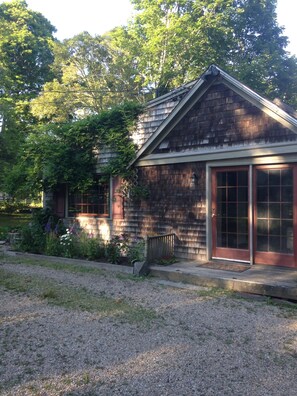 A small wooden building with a red door and windows, surrounded by a gravel driveway and greenery. - This wonderful 6 bedrm 3 bath house & separate cottage is family & dog friendly! (Harwich)