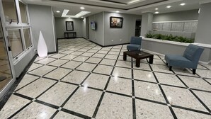 A lobby with a tiled floor, a reception desk, two blue chairs, a small table, and a wall-mounted display.
