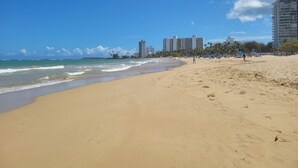 A wide sandy beach with gentle waves, a clear blue sky, and high-rise buildings in the background.