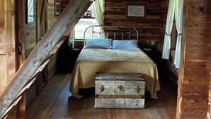 Interior rústico de una cabaña con habitación en altillo, vigas de madera, cama con colcha y un baúl antiguo.