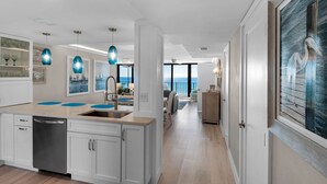 A modern kitchen with white cabinets, a dishwasher, and blue pendant lights. There is a view of the ocean through large windows.