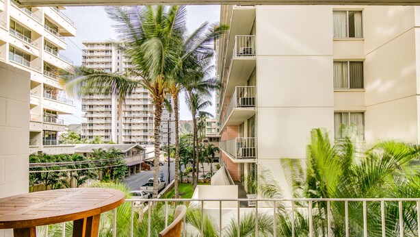 A balcony with a wooden table, surrounded by lush greenery and palm trees, overlooking a street with parked cars and a multi-story building in the background. - Surfer's Dream Condo w/ Full Kitchen - Free Wifi, Sleeps 4 (Honolulu)