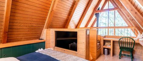 Um quarto de sótão acolhedor com paredes de madeira, teto inclinado, uma cama, um pequeno espaço de estar e uma janela.