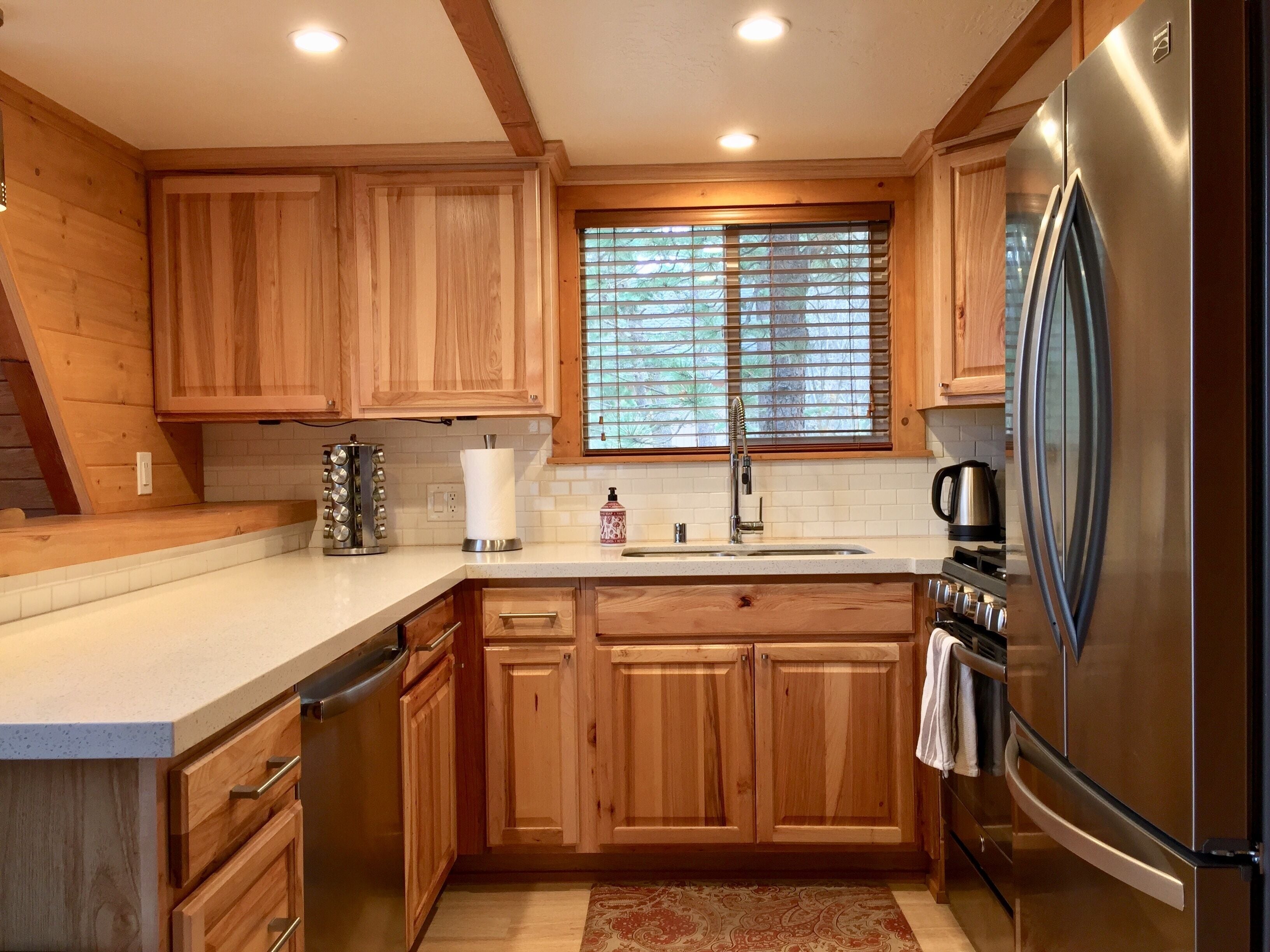 A modern kitchen with wooden cabinets, a stainless steel refrigerator, a sink, and a window with blinds.