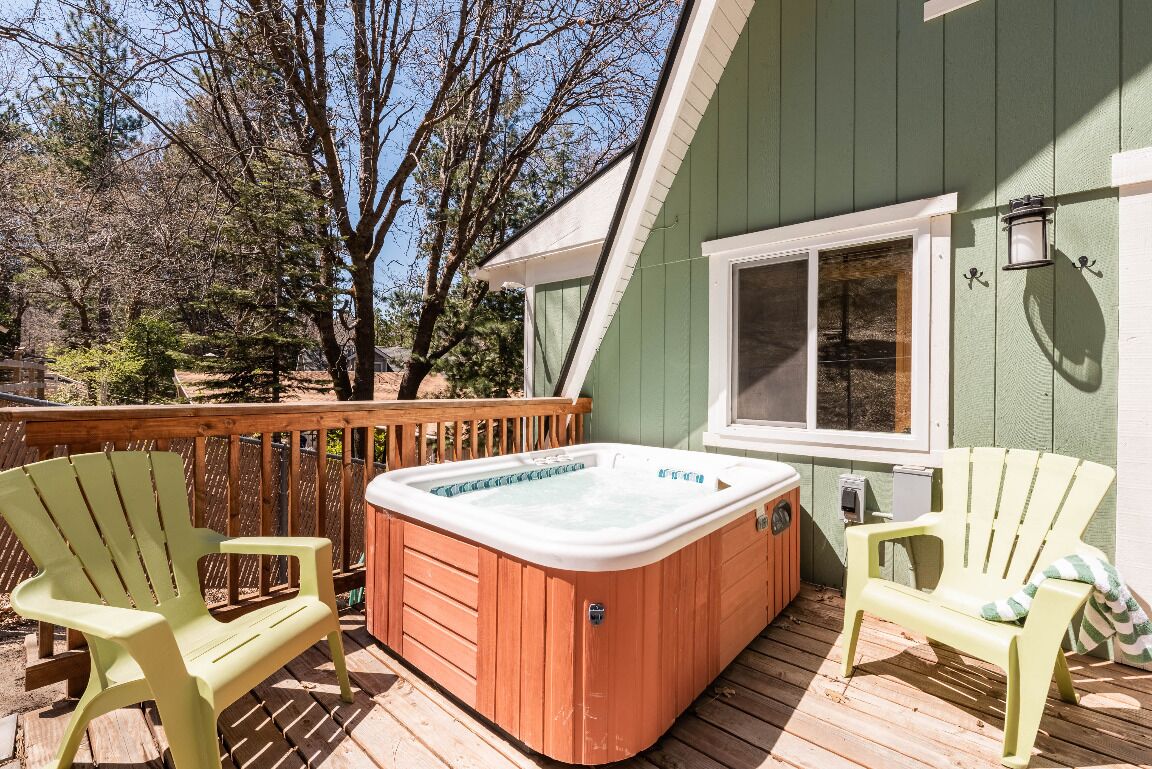 A hot tub on a wooden deck with two Adirondack chairs, surrounded by a wooden railing and a green building.