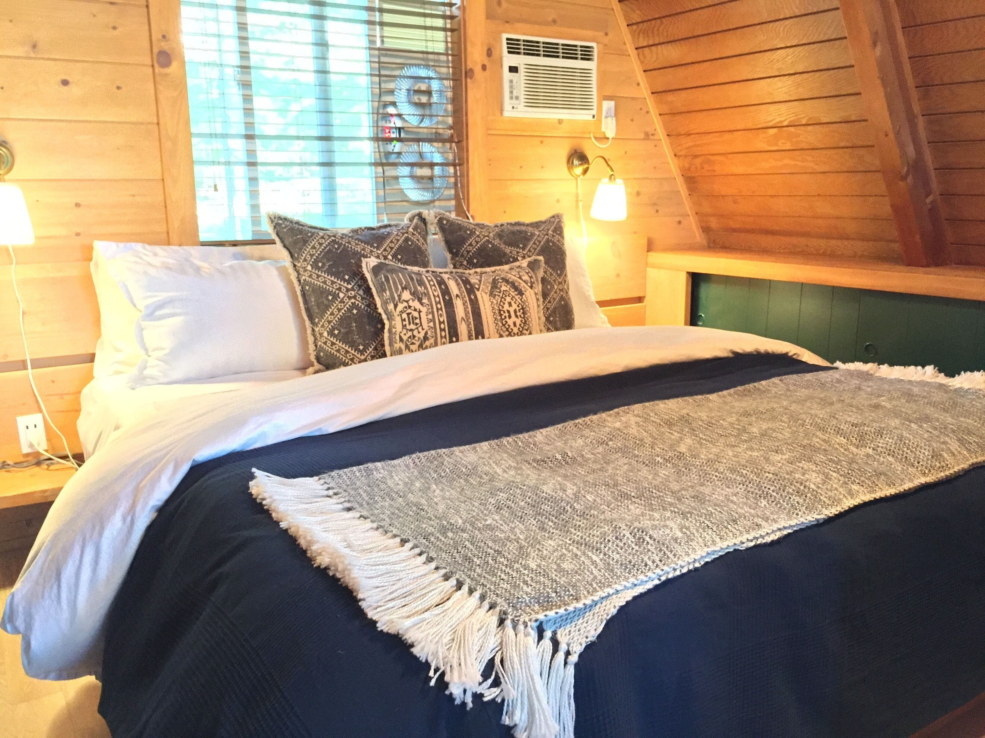 A bedroom with a bed, two bedside lamps, patterned pillows, a woven throw blanket, and a window with blinds.
