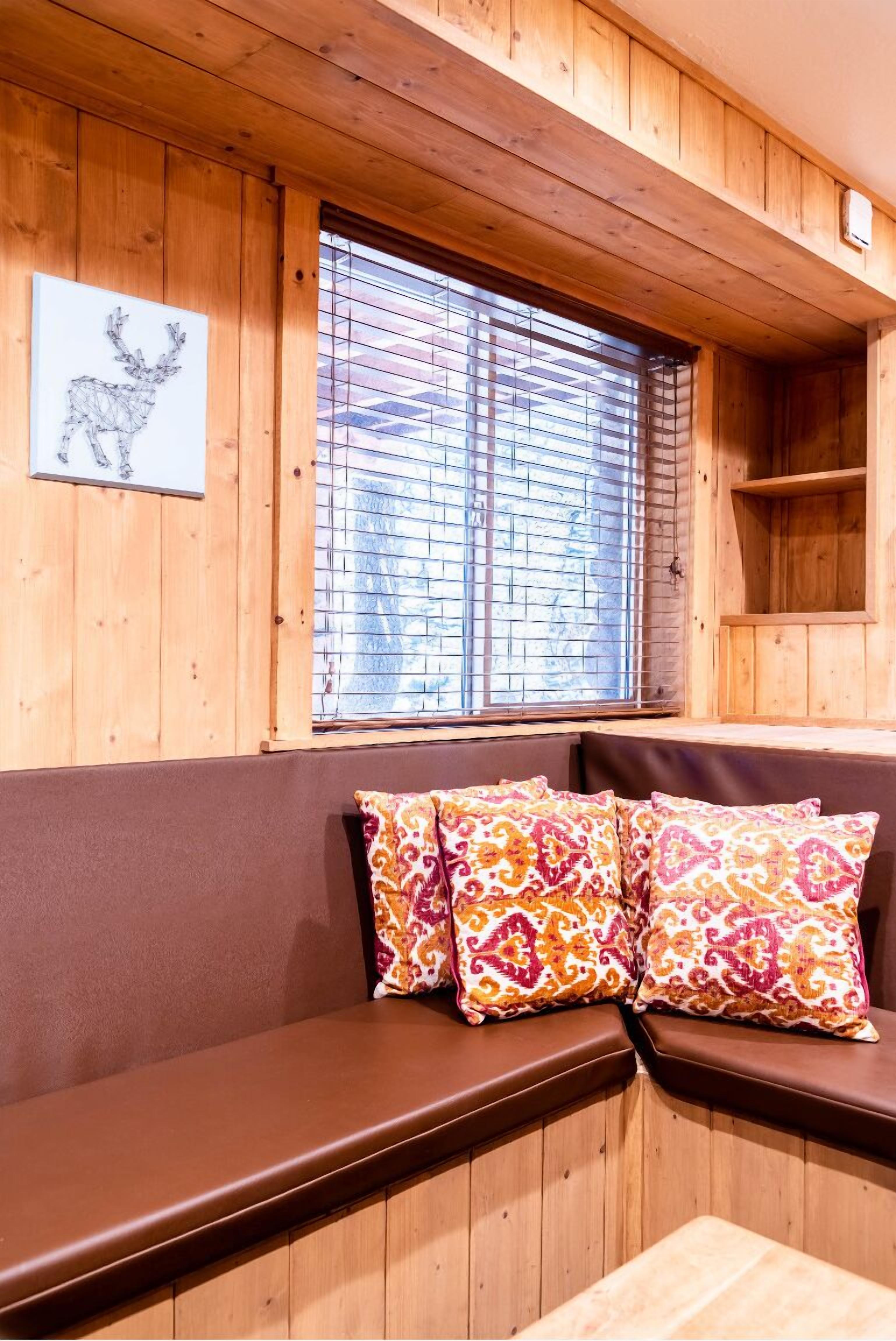 A wooden cabin interior with a bench, patterned cushions, a window with blinds, and a wooden shelf.