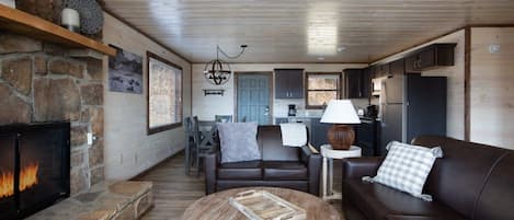 A cozy living room with a fireplace, a sofa, a coffee table, and a kitchen area in the background.