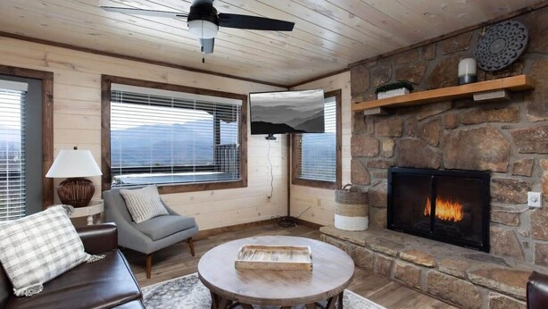 A cozy living room with a stone fireplace, a ceiling fan, and large windows offering a view of the mountains. - Unreal Views GSMNP/Near DwTn Gatlinburg/Hot Tub (Gatlinburg)