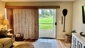 A living room with a sofa, a coffee table, a lamp, a basket, and a view of a green lawn and buildings through a sliding glass door. - Kihei Ground-Floor Condo • Steps to Kamaole III Beach• King Bed • Pool & A/C (Kihei)