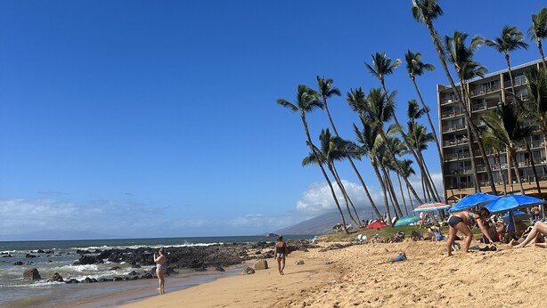 A beach with palm trees, a building with balconies, and people relaxing. - Kihei Ground-Floor Condo • Steps to Kamaole III Beach• King Bed • Pool & A/C (Kihei)