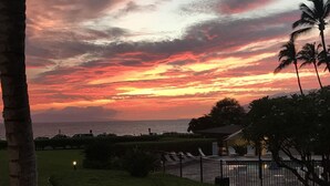 A sunset view over a beach with palm trees and a fenced area with lounge chairs. - Kihei Ground-Floor Condo • Steps to Kamaole III Beach• King Bed • Pool & A/C (Kihei)