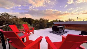 Um terraço no telhado com cadeiras Adirondack vermelhas, uma mesa e vista para o pôr do sol.