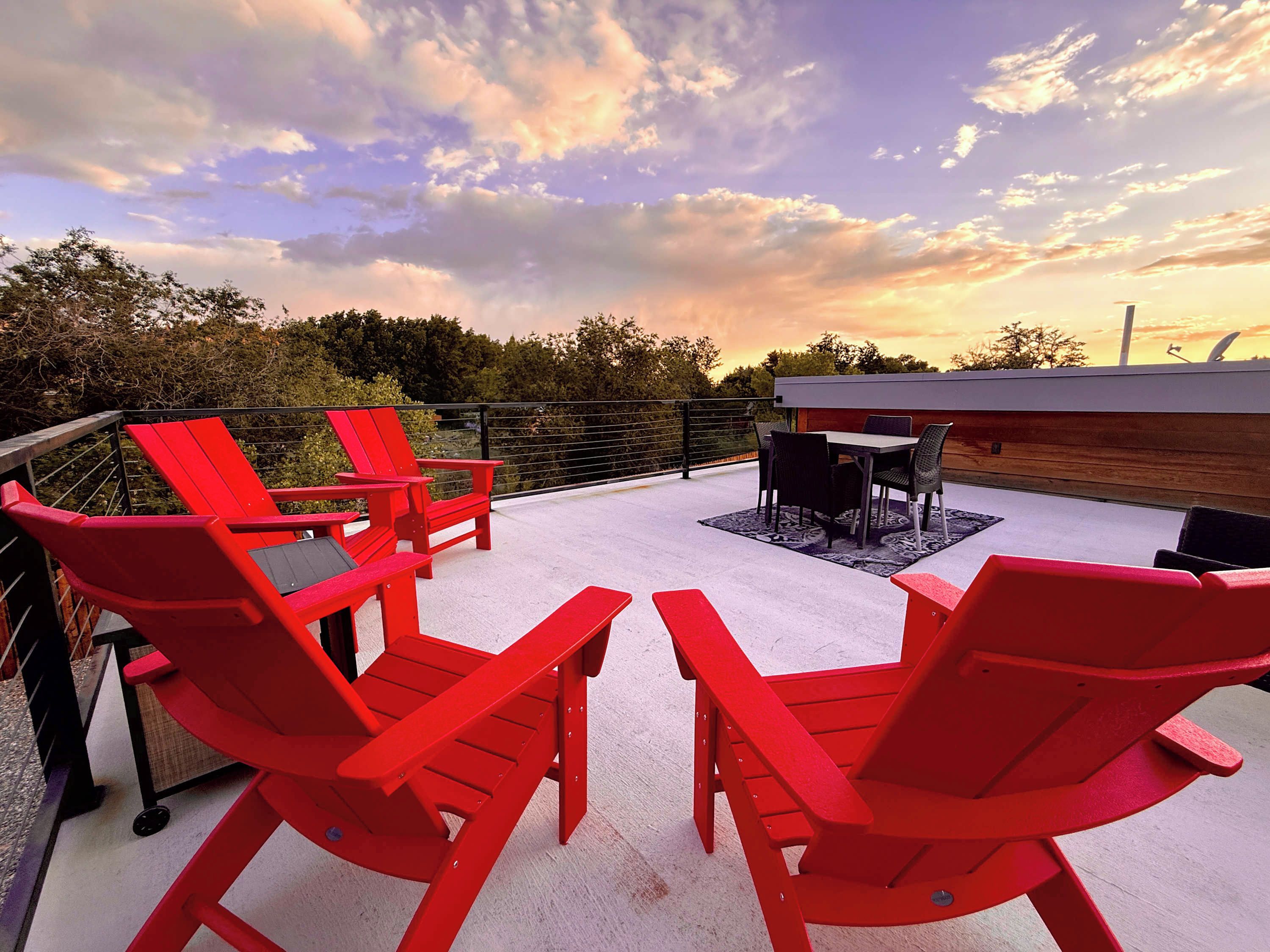 A rooftop patio with red Adirondack chairs, a table, and a view of the sunset.