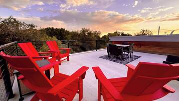 A rooftop patio with red Adirondack chairs, a table, and a view of the sunset.